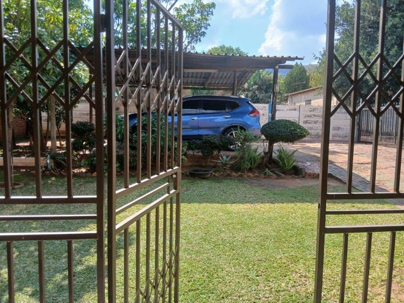 View through a metal security gate towards a front garden, paved driveway, and a covered carport structure.