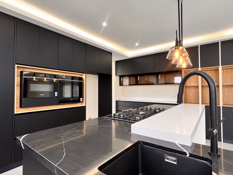 Modern kitchen featuring matte black cabinetry, stone countertops, a central island with a gas hob, and integrated high-end appliances with ambient cove lighting.
