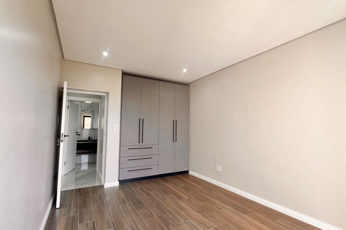 A clean, modern bedroom featuring neutral tones, wood-look tiled flooring, and ample built-in cupboard space, with direct access to an en-suite bathroom.
