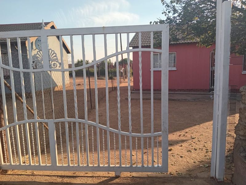 Front view of a residential property featuring a white metal security gate, wire fencing, and a red exterior facade with tiled roofing.