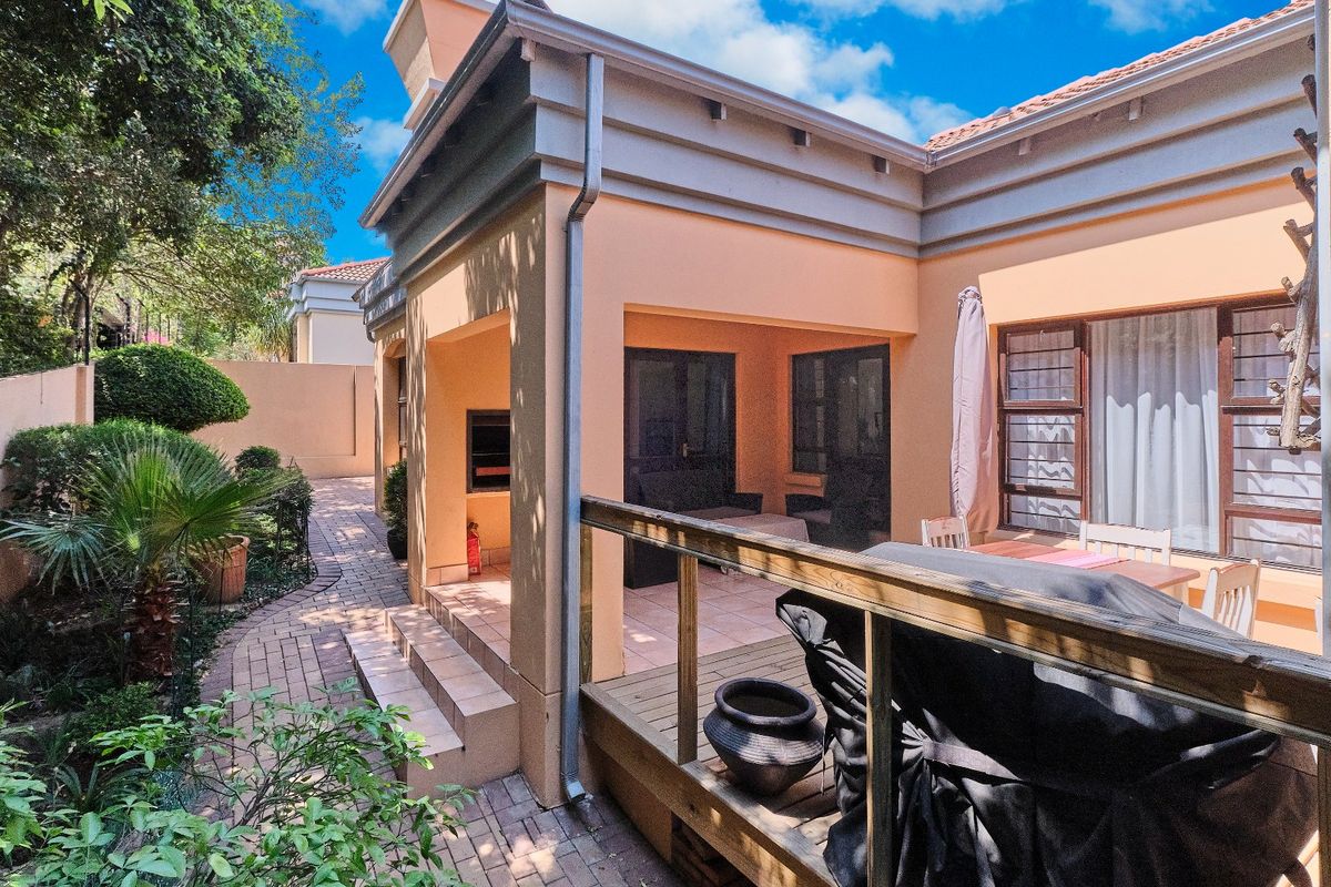 A well-maintained rear exterior featuring a covered patio with a built-in braai, adjacent timber decking, and a paved garden walkway.