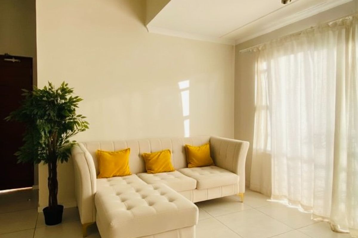 A bright living room featuring neutral tiled flooring, a recessed ceiling design with spotlighting, and large windows dressed with sheer curtains.