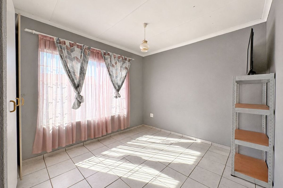 A simple, tiled bedroom featuring a large window with security bars and neutral grey walls, offering a clean and functional space.