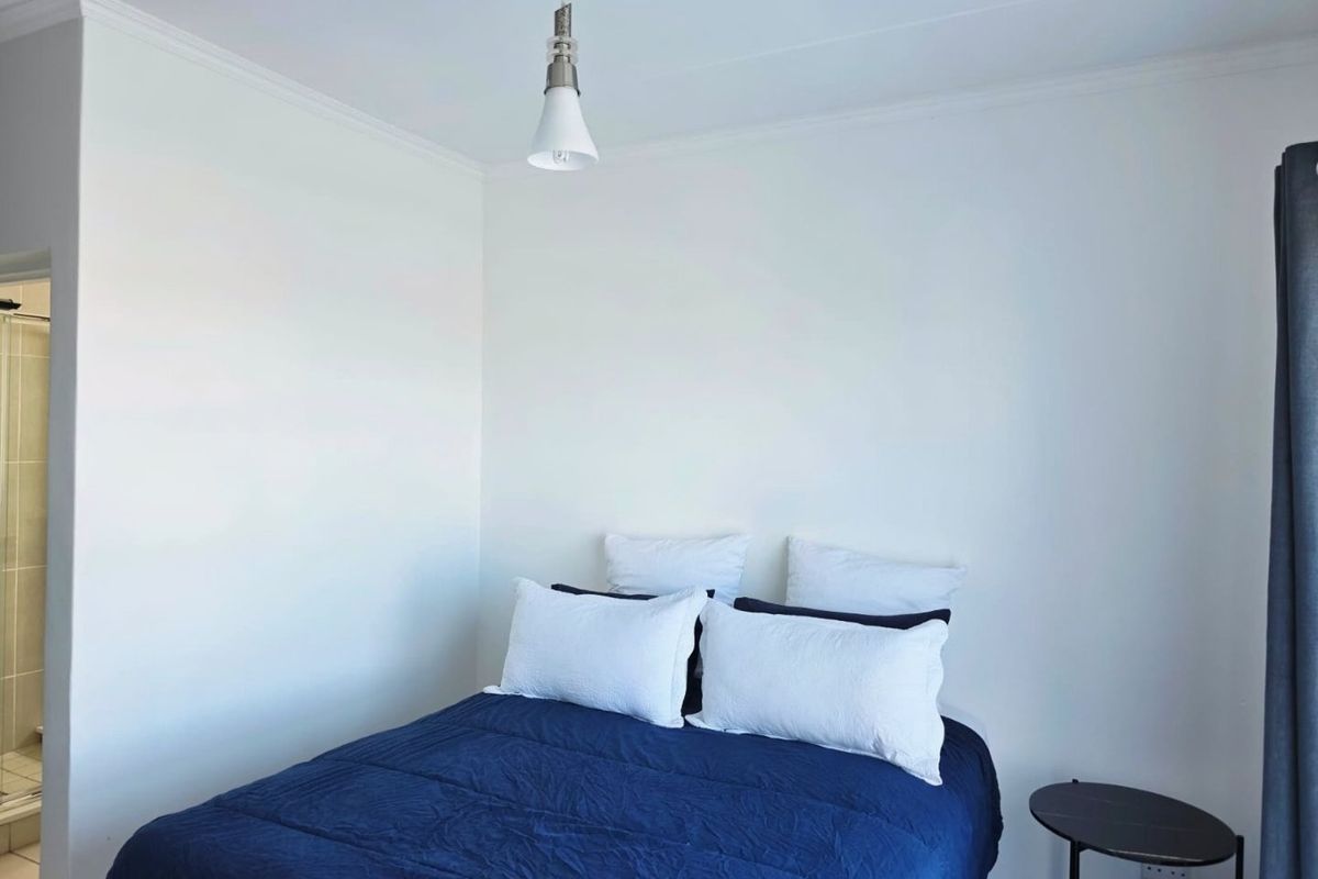 A simple bedroom featuring white walls, coving, and a pendant light fixture, with an adjacent bathroom visible.