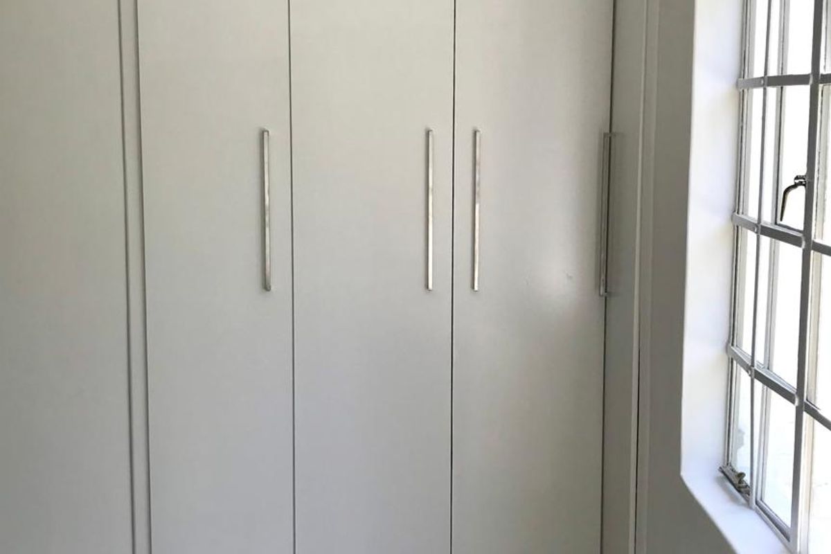 A bright, empty bedroom featuring a clean white built-in wardrobe, dark laminate flooring, and a classic steel-framed window.