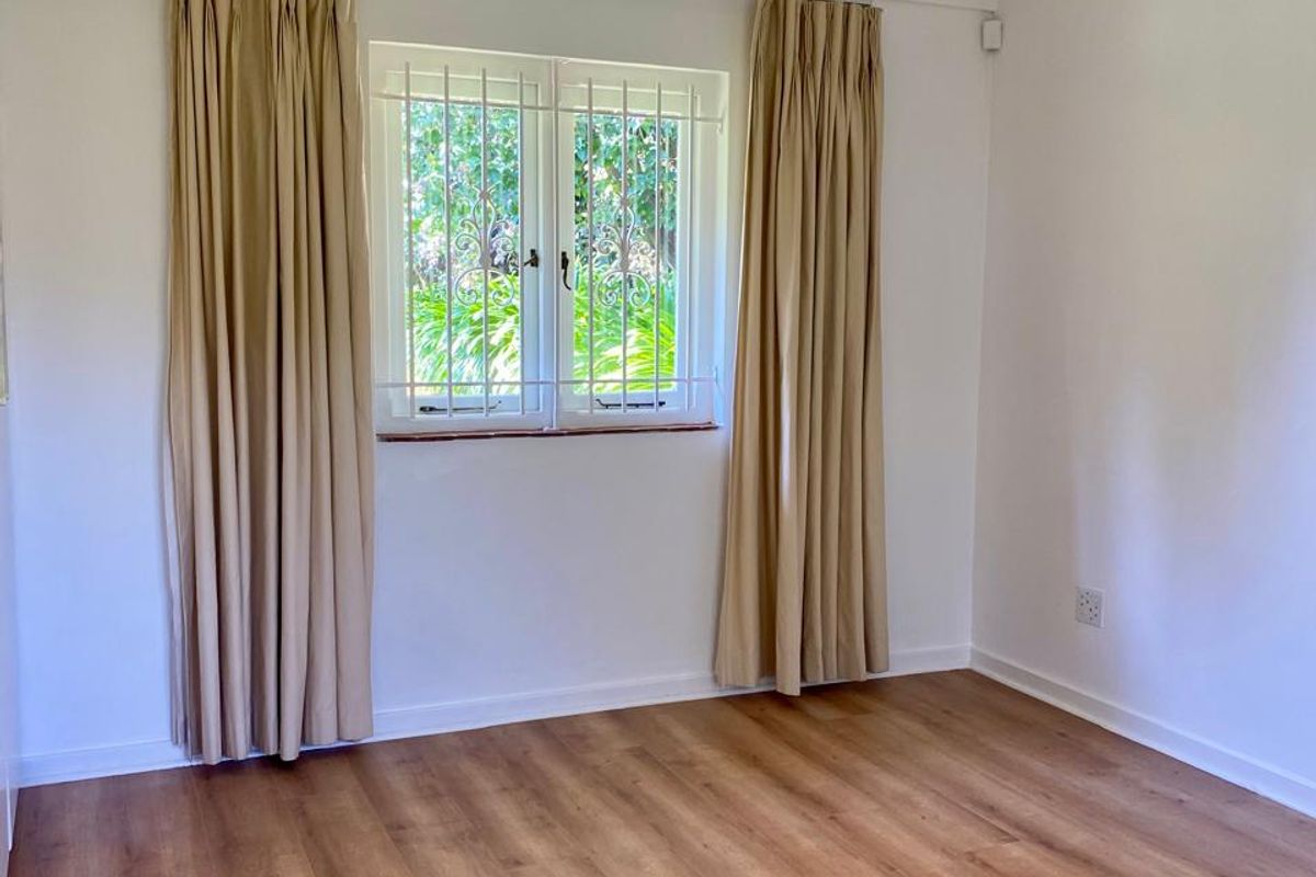 A bright, empty bedroom featuring modern laminate flooring, recessed downlights, and a window fitted with security bars.