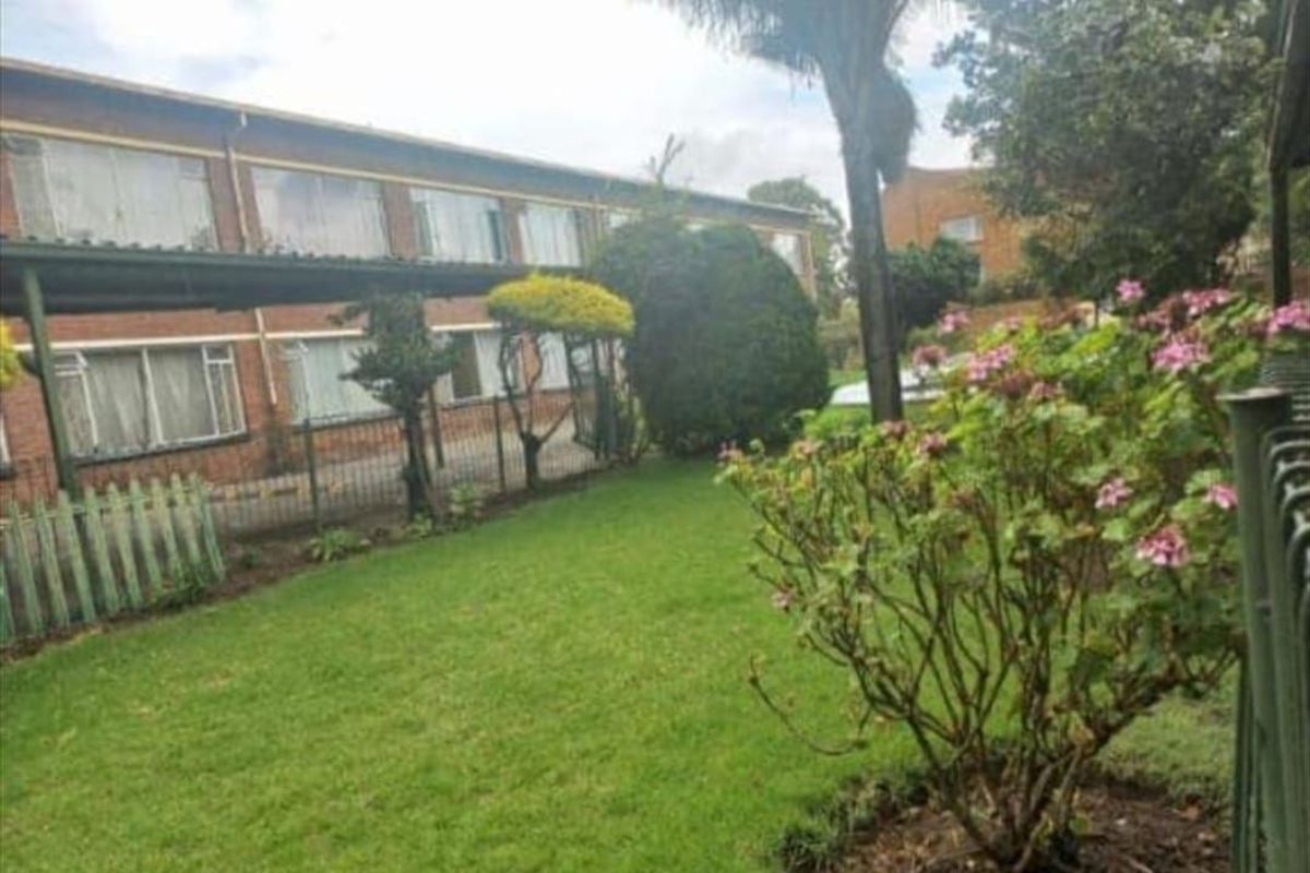 A view of the rear exterior of the apartment building, featuring a well-maintained lawn, established garden beds, and a covered walkway.