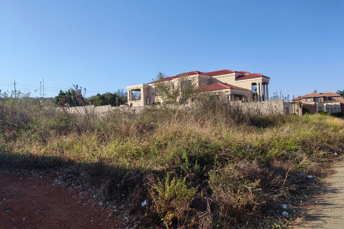A view of a vacant residential plot with overgrown vegetation, featuring a perimeter wall with electric fencing and a neighboring house in the background.