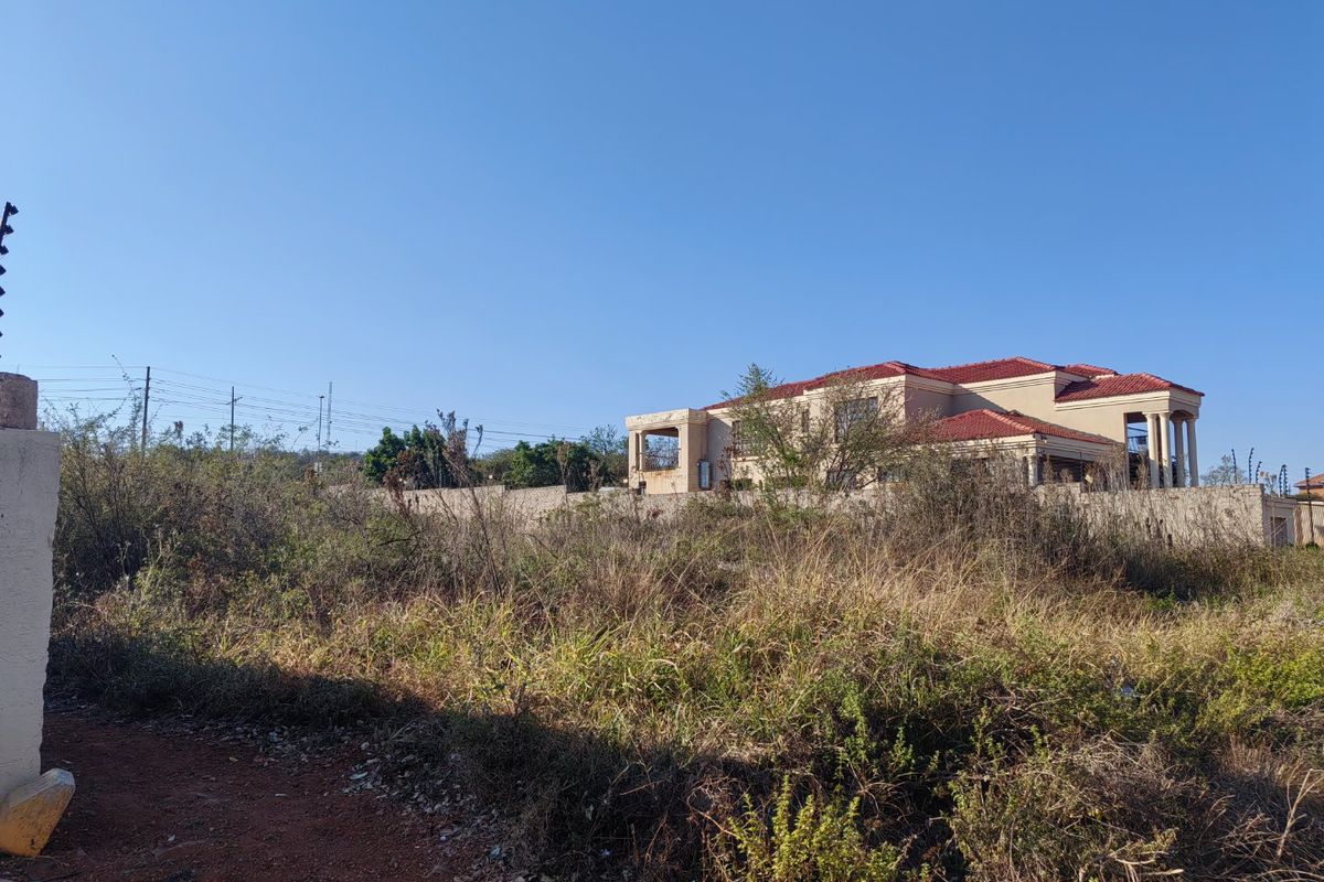 A vacant residential plot featuring overgrown vegetation, enclosed by a perimeter wall with electric fencing, offering a view of neighboring properties.