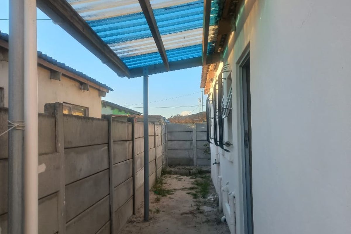 A narrow rear exterior passage featuring a corrugated plastic lean-to roof, precast concrete boundary wall, and a concrete pathway leading to an external door.