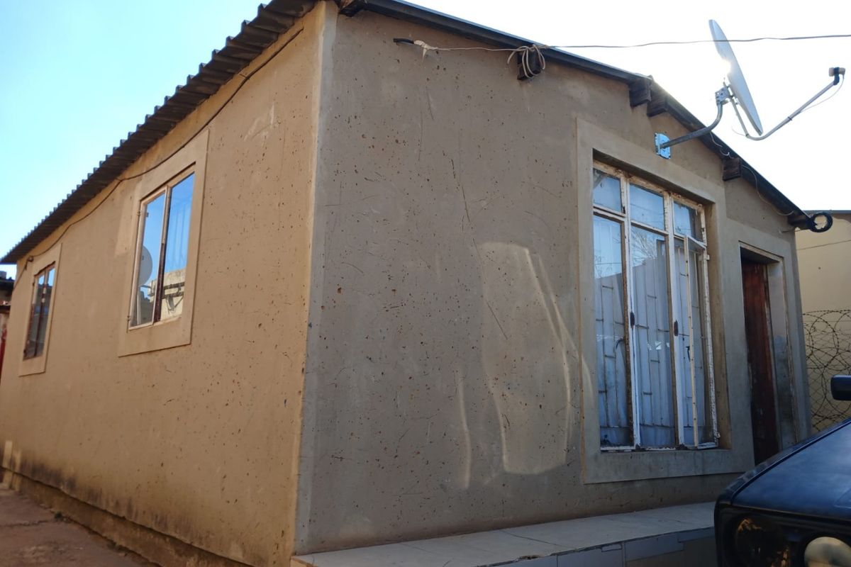 Exterior view of a single-story residential structure featuring a corrugated metal roof, metal-framed windows with security bars, and a tiled entrance step.