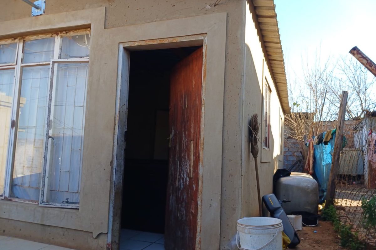 Rear exterior view of a modest residential structure featuring plastered walls, a timber entrance door, and aluminium-framed windows with security bars.