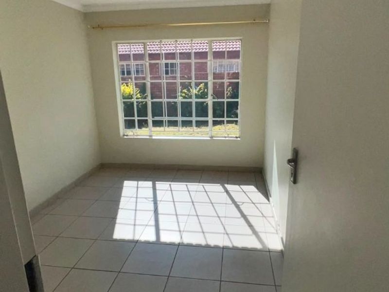 A bright, empty bedroom featuring neutral tiled flooring, a large window with security bars, and a simple ceiling light fixture.