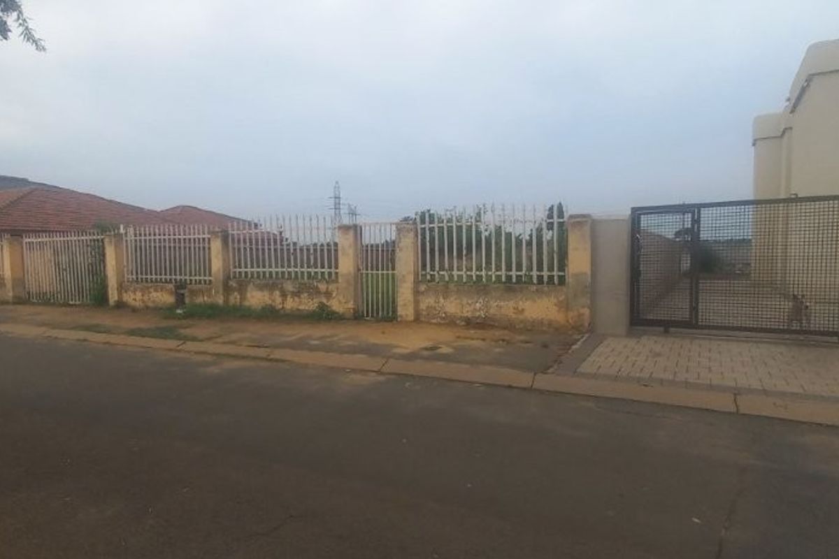 Street view of a residential vacant plot featuring a perimeter wall, metal security fencing, and a gated paved driveway entrance.