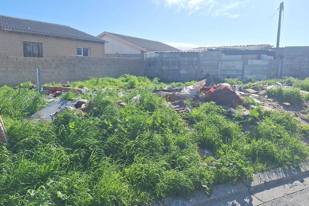 Vacant residential plot enclosed by concrete block boundary walls, currently overgrown with vegetation and containing accumulated debris.