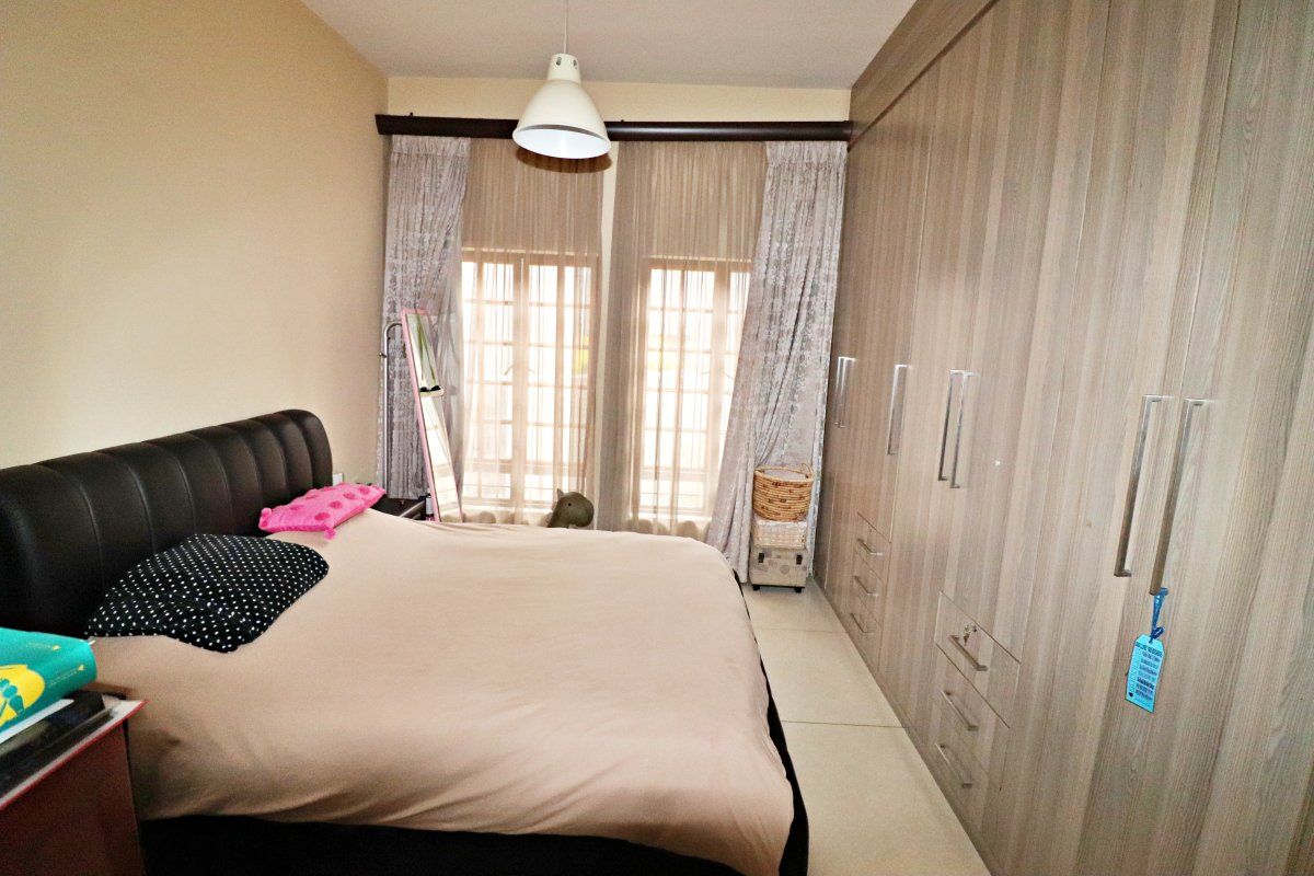 A well-maintained bedroom featuring extensive built-in wooden cupboards, tiled flooring, and large windows with curtain rails for natural light.