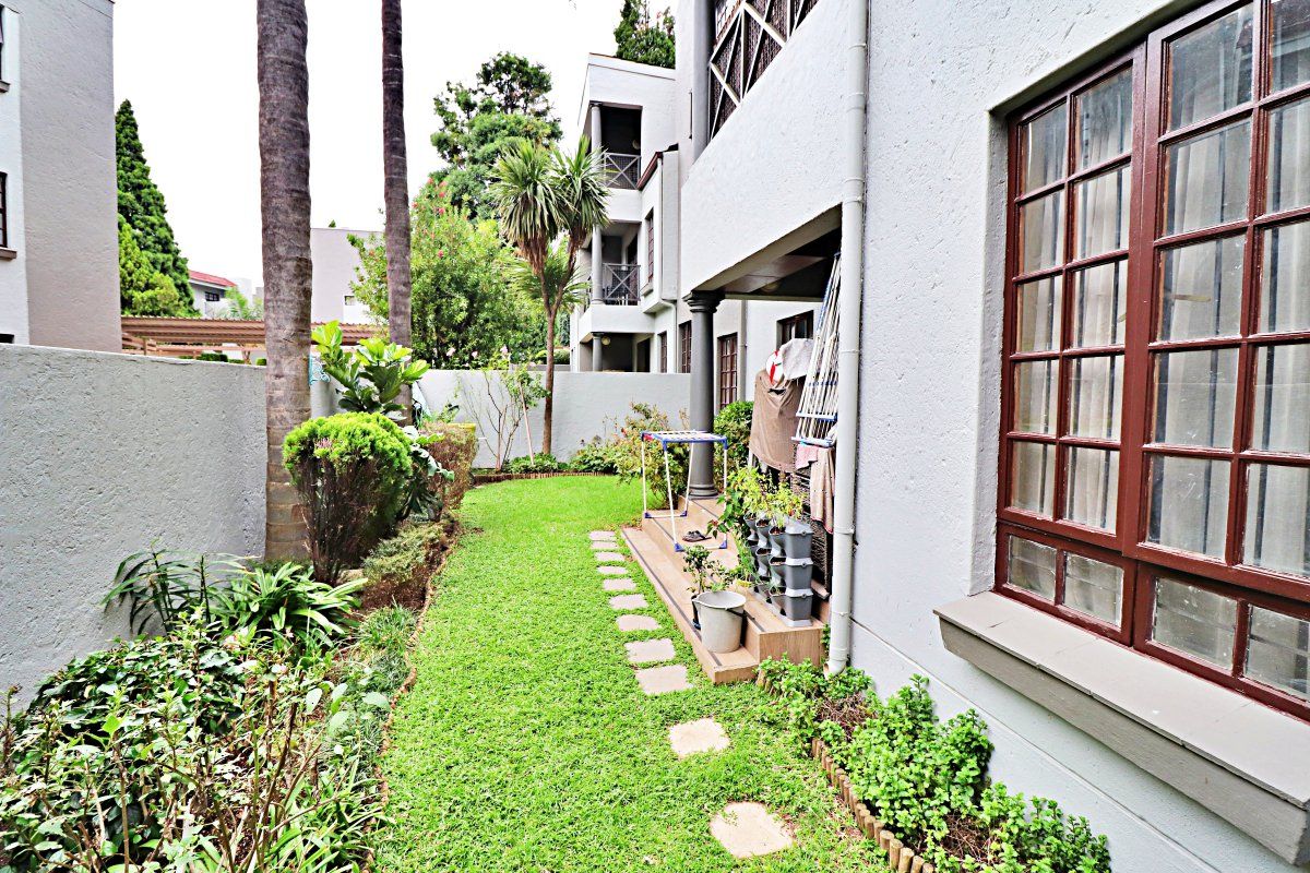A private rear garden featuring a manicured lawn, stone pathway, and established landscaping, adjacent to the apartment's covered patio and exterior facade.