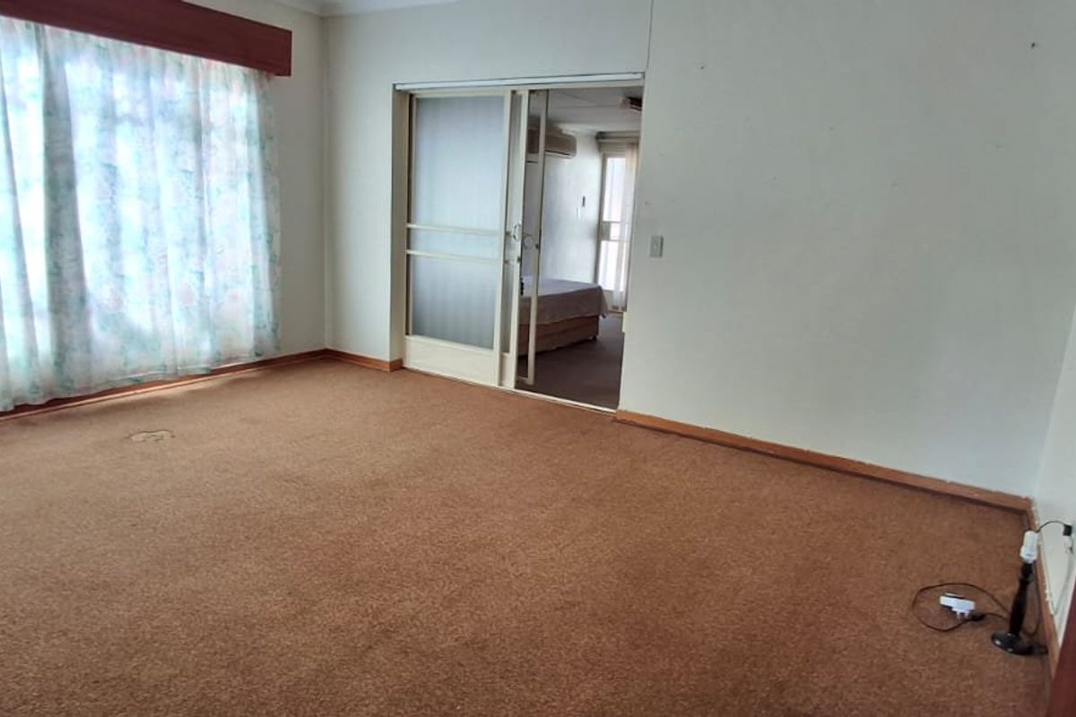 A spacious, empty bedroom featuring carpeted flooring, a large window with a pelmet, and a sliding door leading to an adjacent room.