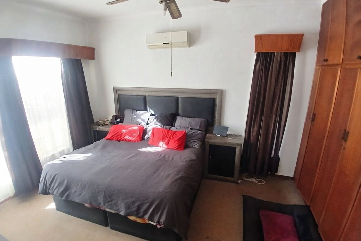 A bedroom featuring a built-in wooden wardrobe, ceiling fan, and wall-mounted air conditioning unit, with carpeted flooring and window pelmets.