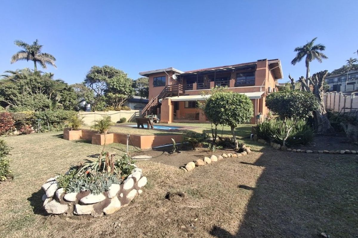 Rear view of the property featuring a swimming pool, covered patio, upper balcony, and a landscaped garden with stone retaining walls.