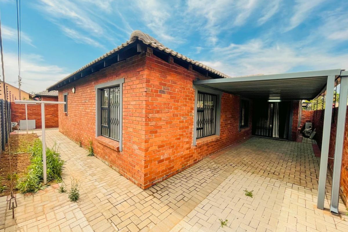 Rear view of a face brick house featuring a paved driveway, covered carport, and secure window bars.