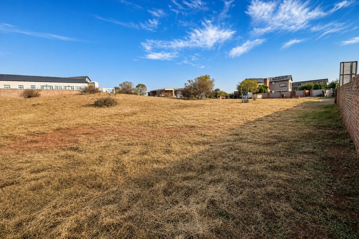 A vacant residential plot featuring dry grass and a brick boundary wall, situated within a developed neighborhood.