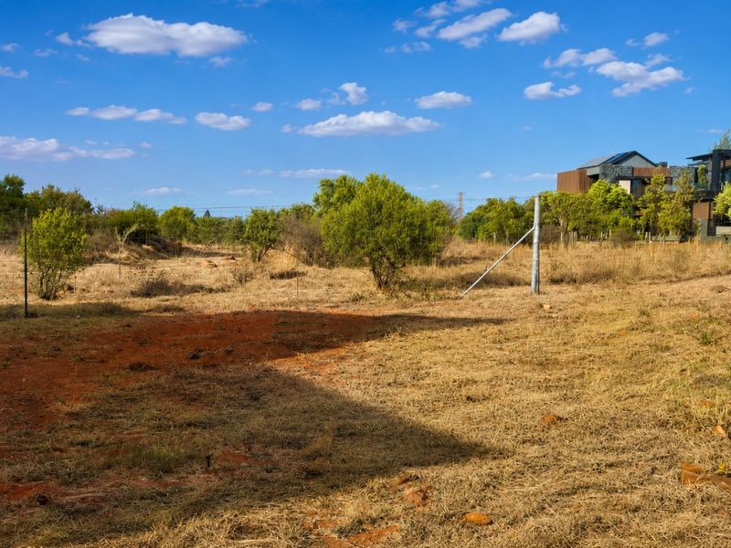 A view of a vacant residential plot featuring dry grass, native shrubs, and perimeter wire fencing with metal posts.