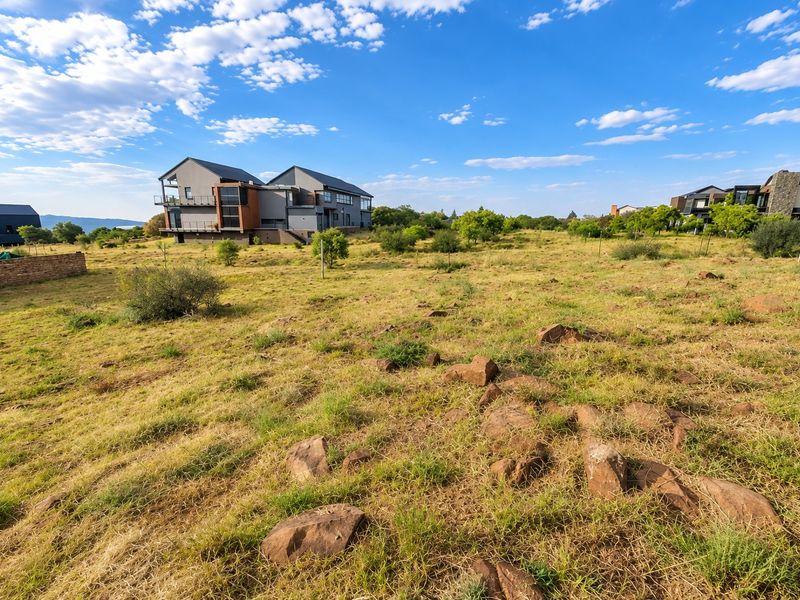 A view of a vacant residential plot featuring natural rocky terrain and native vegetation, with neighboring modern homes visible in the background.