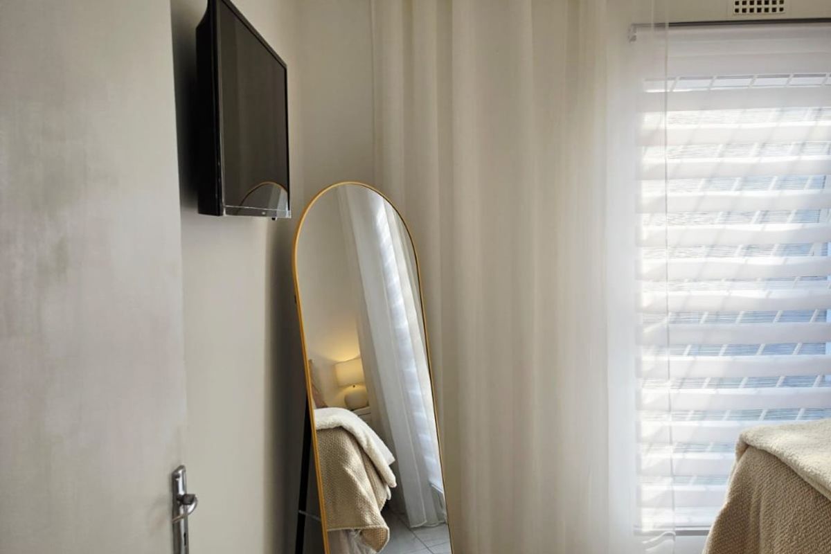 A bright bedroom featuring polished tiled flooring, a wall-mounted television, and large windows dressed with sheer curtains.
