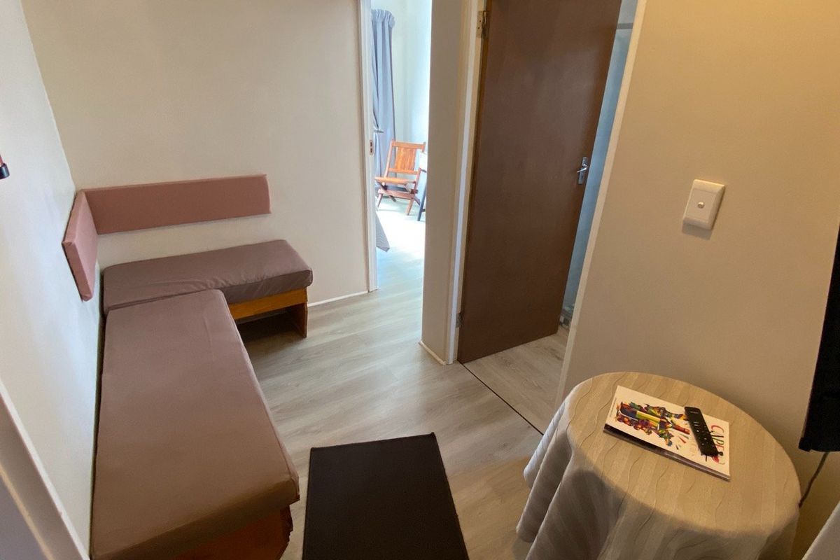 A compact bedroom featuring light-toned laminate flooring, built-in bench seating, and access to an adjoining room through an internal doorway.