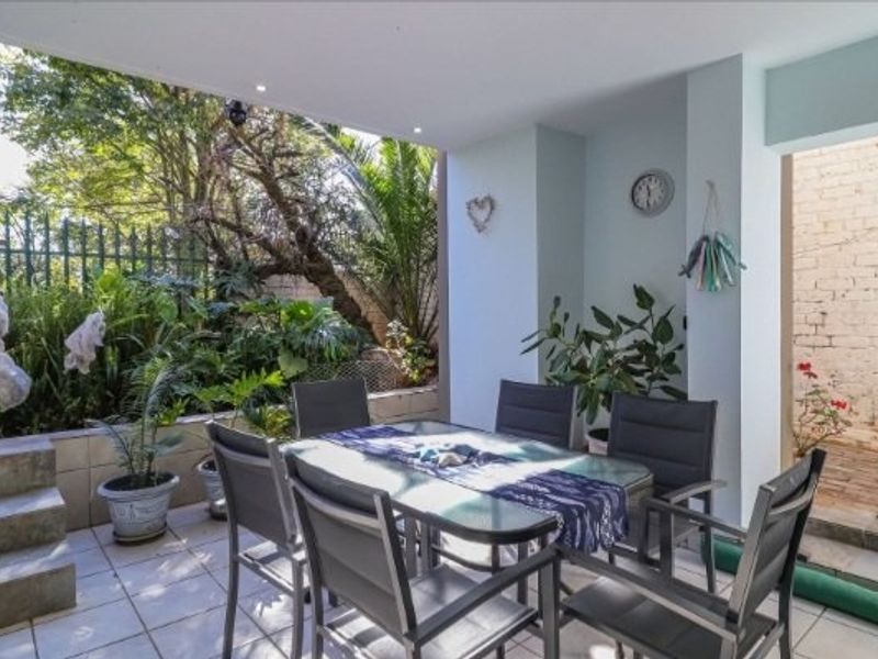 A covered outdoor patio area featuring tiled flooring, recessed lighting, and direct access to a garden space with a brick retaining wall.