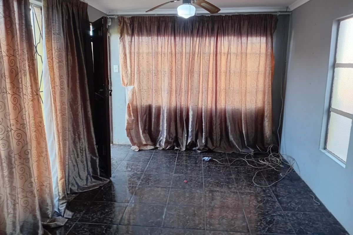 An empty living room featuring dark tiled flooring, a ceiling fan, and large windows covered by curtains.