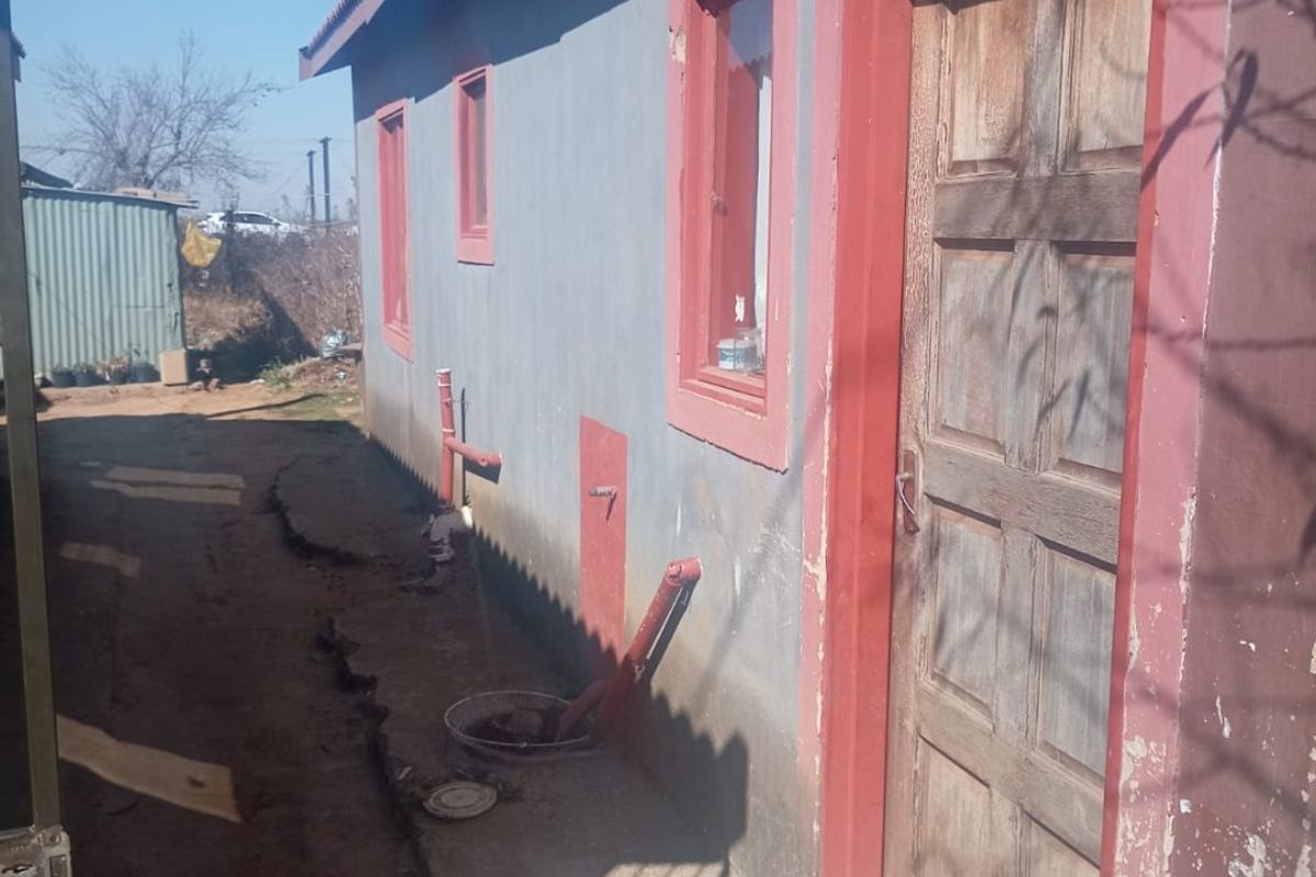Rear exterior view of the property showing a concrete pathway, timber door, and visible signs of foundation cracking and exterior wall wear.