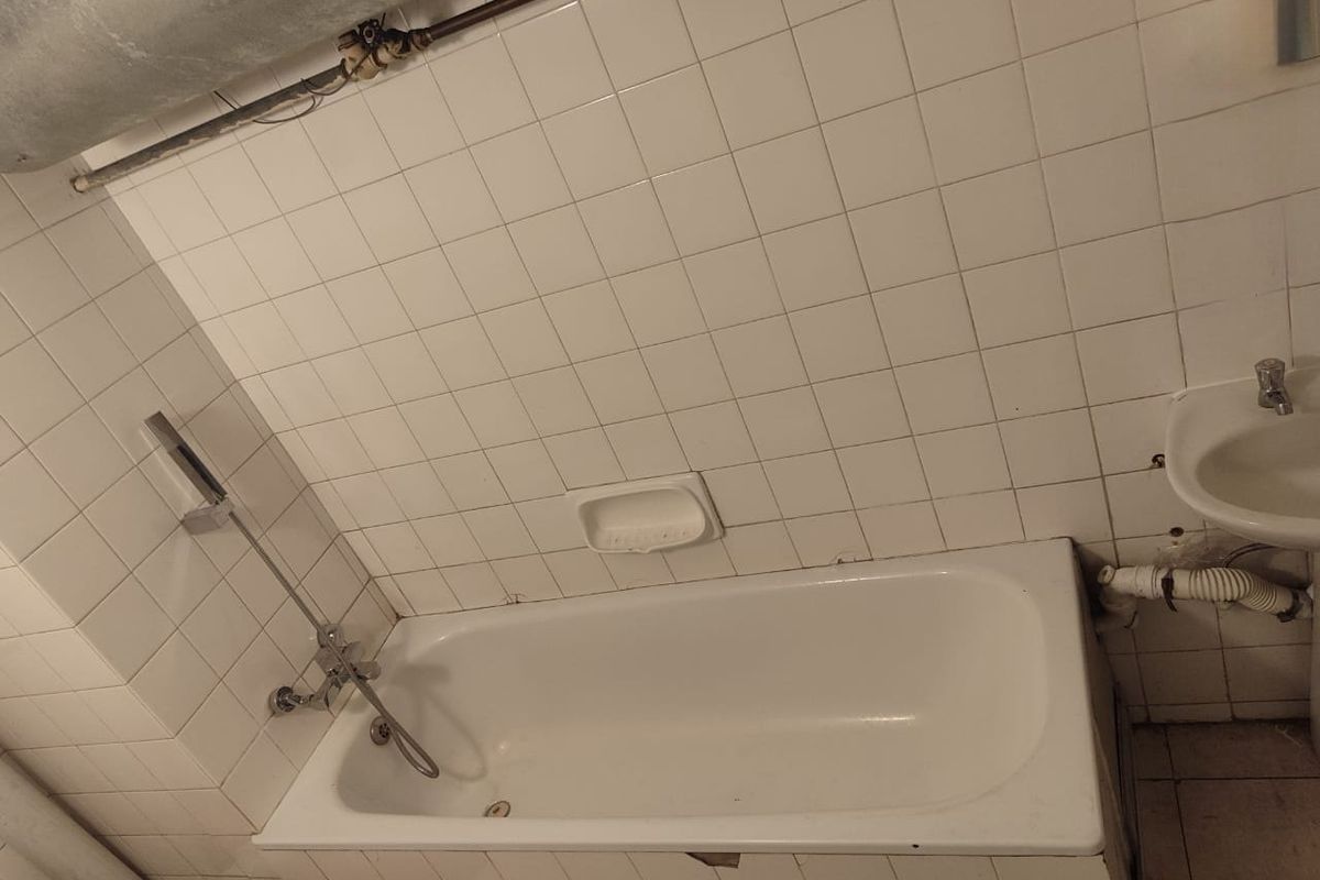 A dated bathroom featuring a standard bathtub, wall-mounted shower mixer, and pedestal basin, requiring renovation to address exposed plumbing and damaged tile finishes.