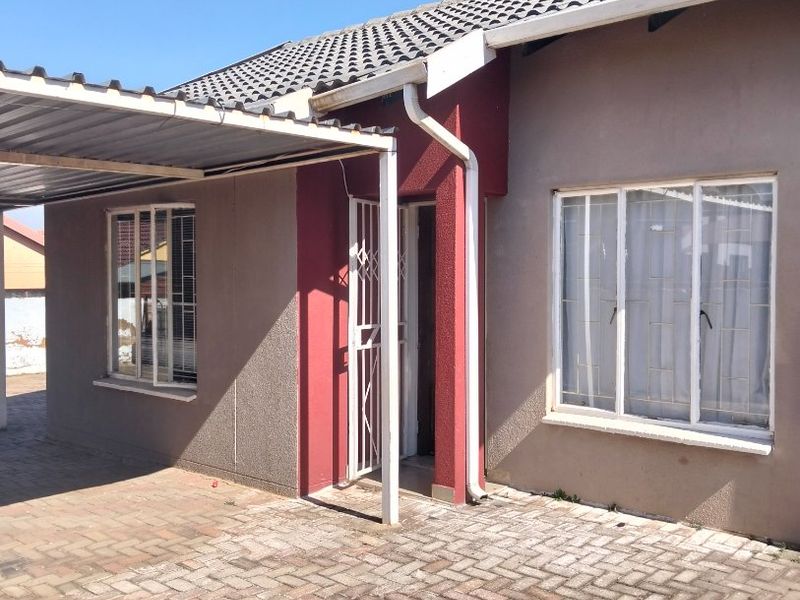 Front elevation of a single-story house featuring a paved driveway, attached carport, and security features including burglar bars and a security gate.