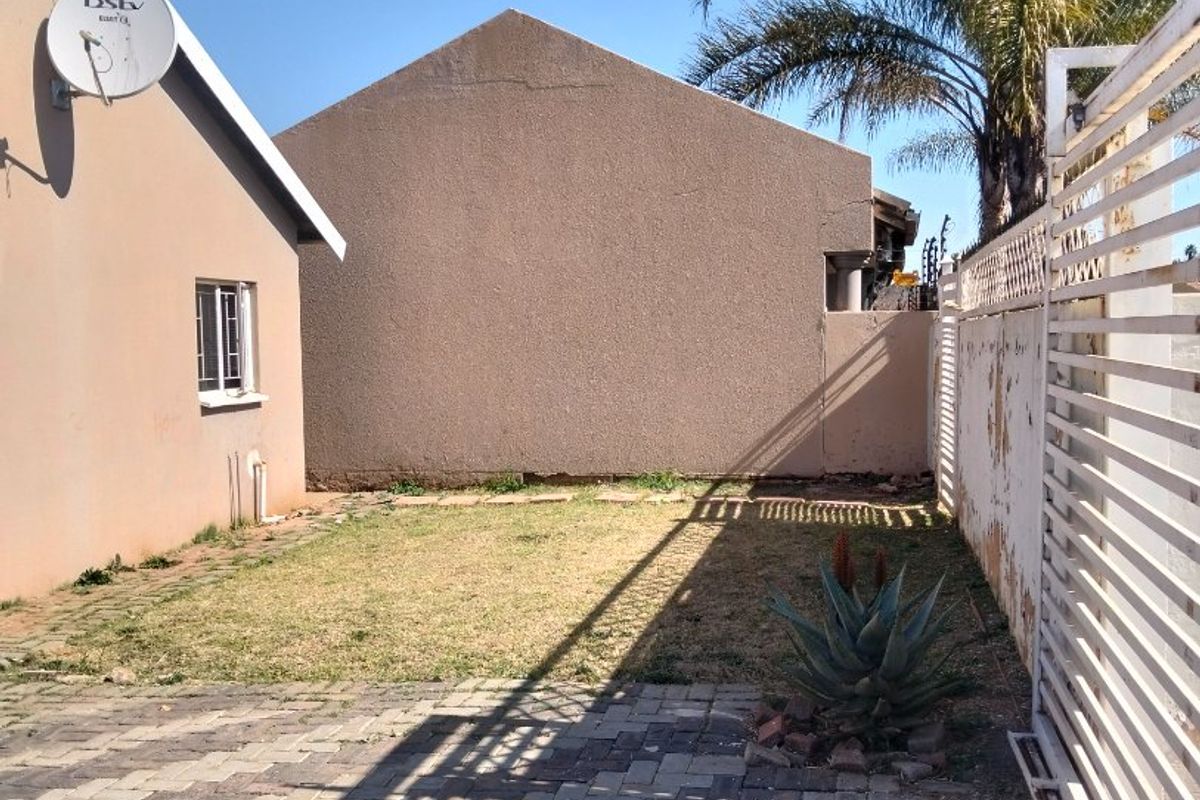 A rear exterior view showing a paved area, a small lawn, and a boundary wall with visible wear and peeling paint.