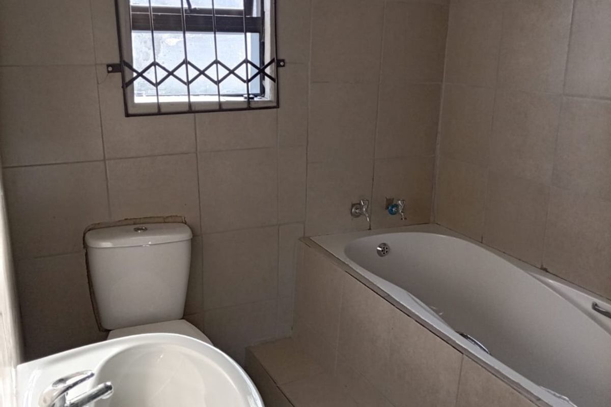 A functional bathroom featuring a pedestal basin, toilet, and a tiled built-in bathtub with a security-gated window.
