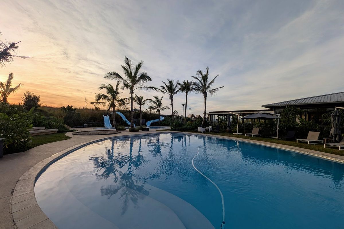 A well-maintained swimming pool area featuring a slide, paved decking, and lush landscaping, set against a scenic sunset backdrop.