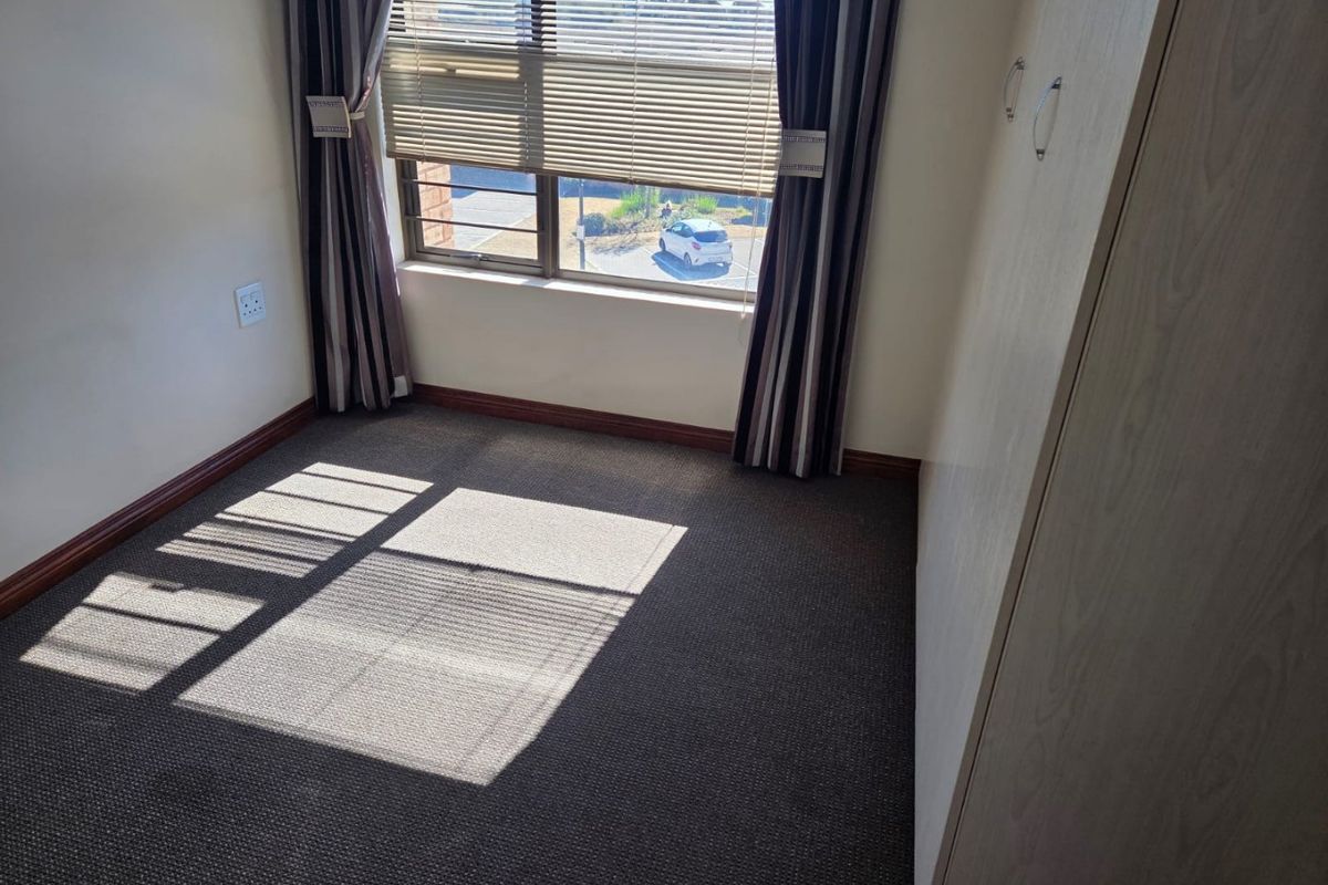 A compact, empty bedroom featuring neutral carpeted flooring, a built-in wardrobe, and a window with venetian blinds providing natural light.