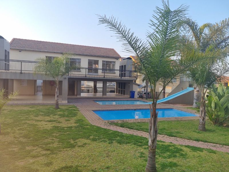 A communal outdoor area featuring a swimming pool with a slide, paved surround, landscaped garden, and a raised balcony overlooking the pool.