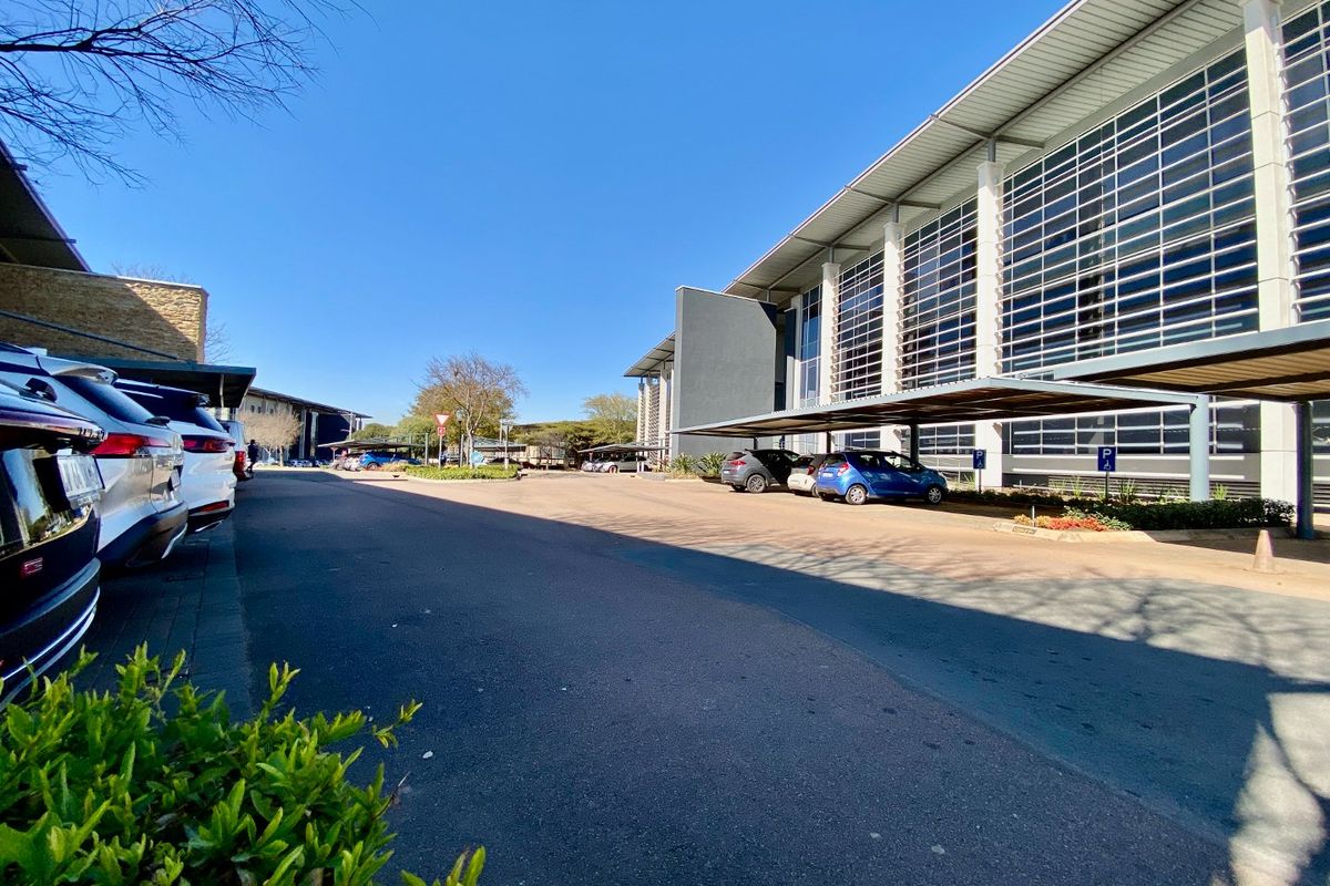 Modern commercial office building exterior featuring extensive glass curtain walling, covered parking bays, and well-maintained asphalt driveway access.