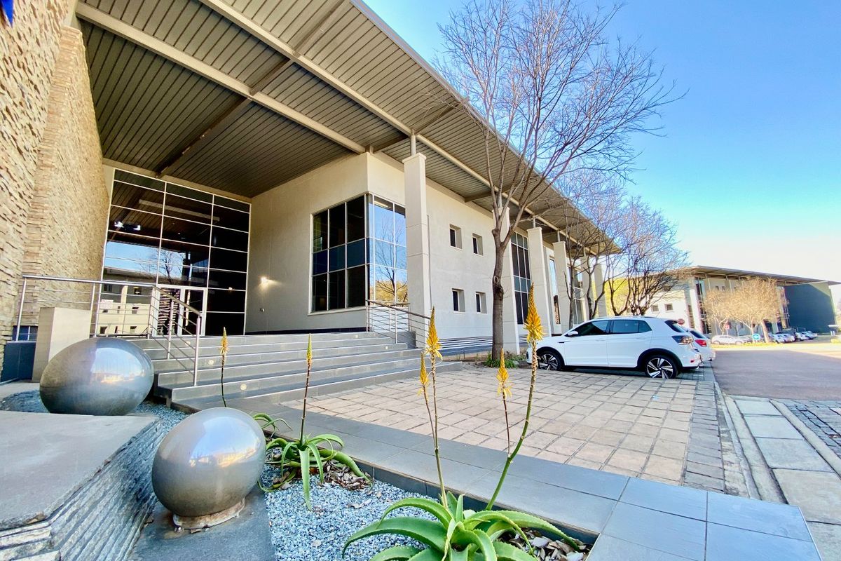 Modern commercial building exterior featuring a prominent metal roof, glass facade, stone cladding, and paved entrance area with stainless steel handrails.
