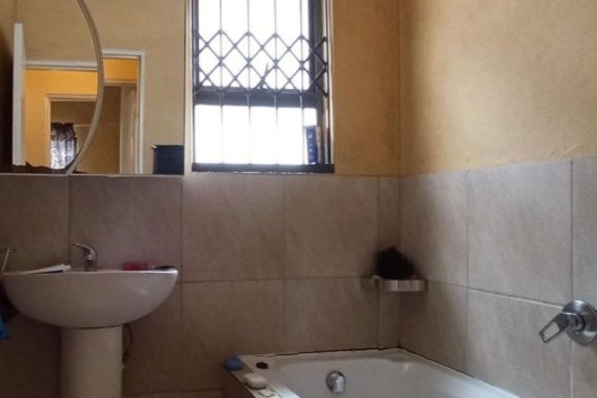 A functional bathroom featuring a pedestal basin, bathtub with mixer tap, and security bars on the window, finished with neutral wall tiling.