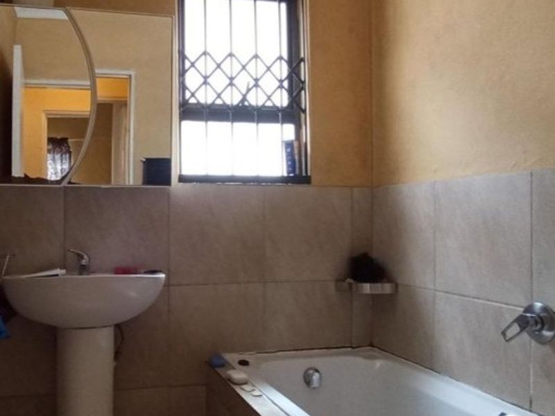 A functional bathroom featuring a pedestal basin, bathtub with mixer tap, and security bars on the window, finished with neutral wall tiling.