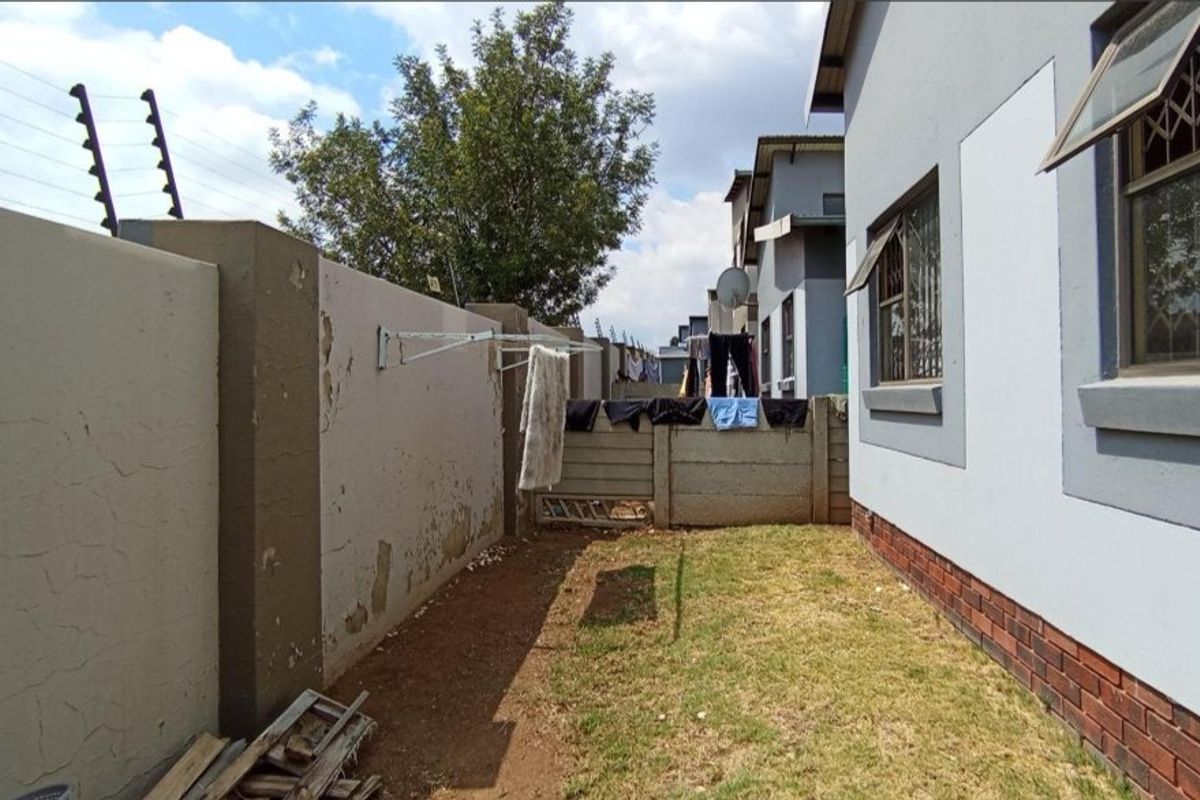 Rear exterior view of the townhouse featuring a small lawn area, perimeter wall with electric fencing, and brick-based exterior wall finish.