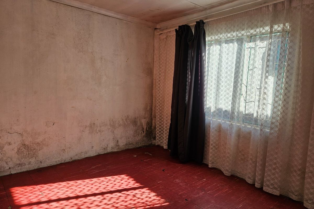 An empty bedroom featuring red tiled flooring and security bars on the window, requiring significant wall repairs and cosmetic updates.