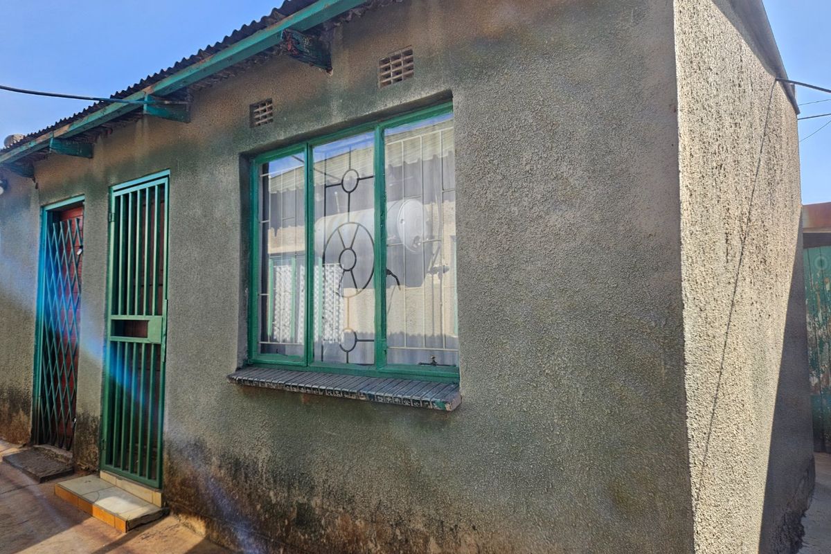 Exterior view of the property showing a plastered wall, metal window frames with security bars, and a corrugated metal roof.