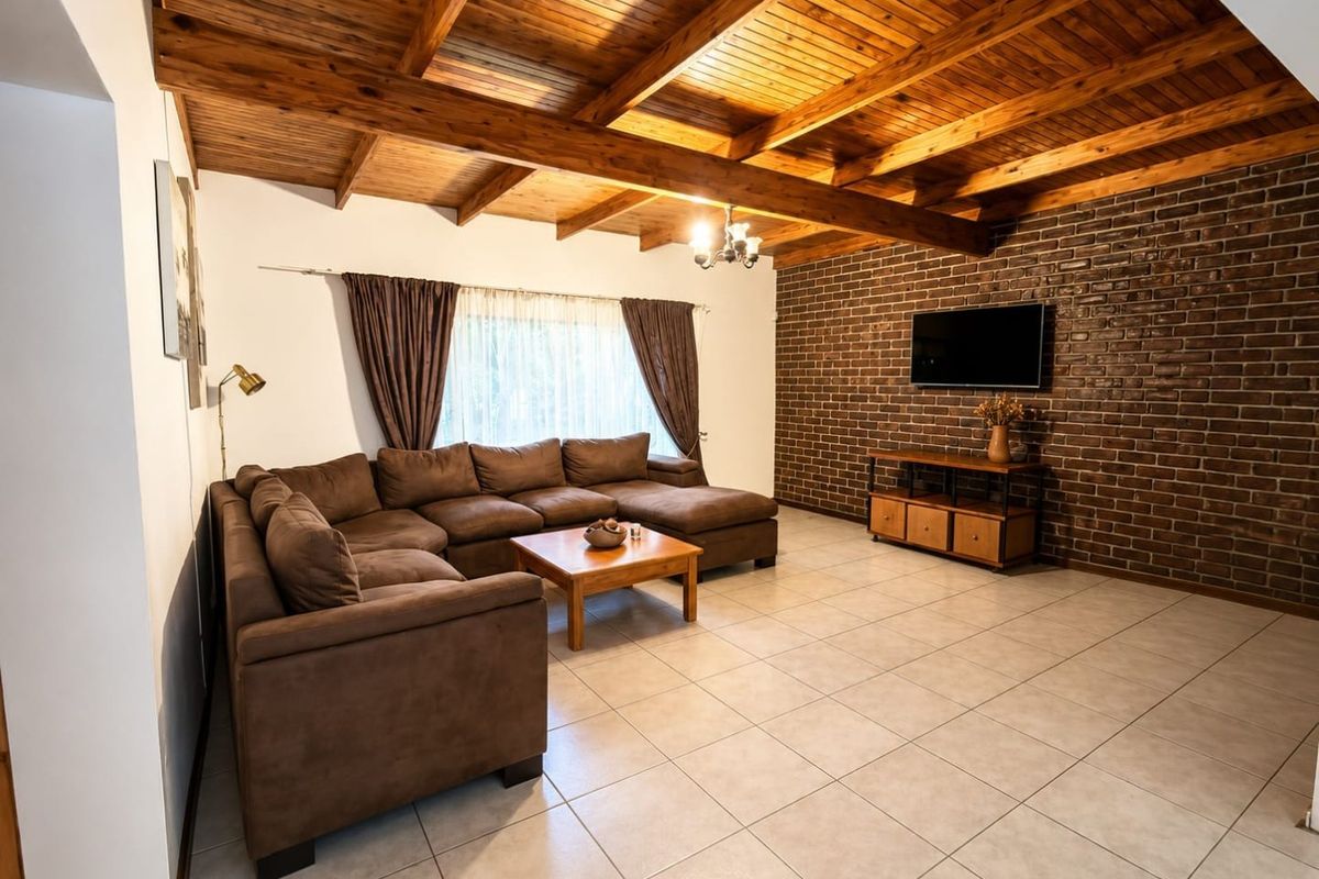 Spacious living room featuring a high exposed timber beam ceiling, a rustic exposed brick feature wall, and neutral ceramic floor tiles.