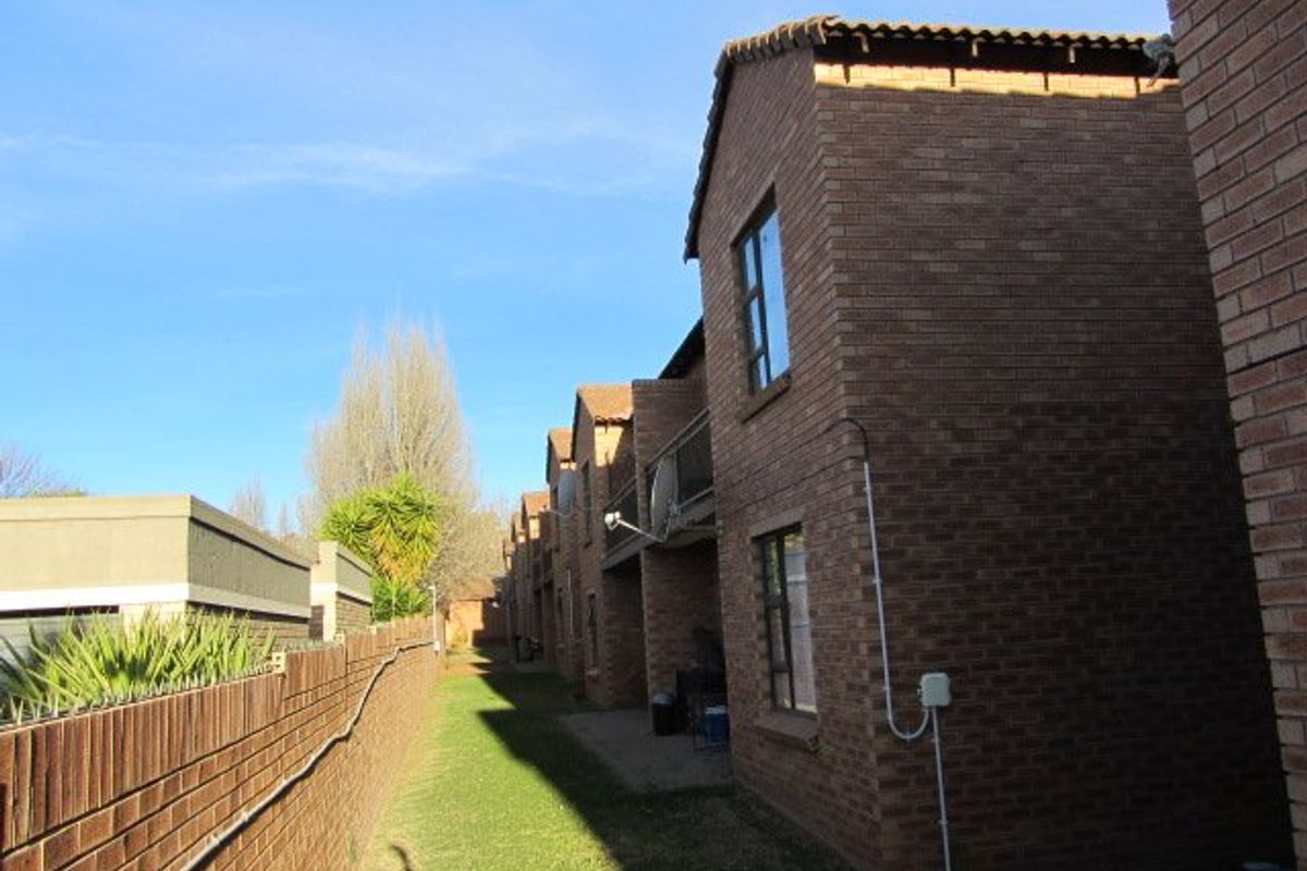 Rear view of a face brick apartment complex featuring a paved walkway, landscaped lawn, and perimeter wall with security spikes.