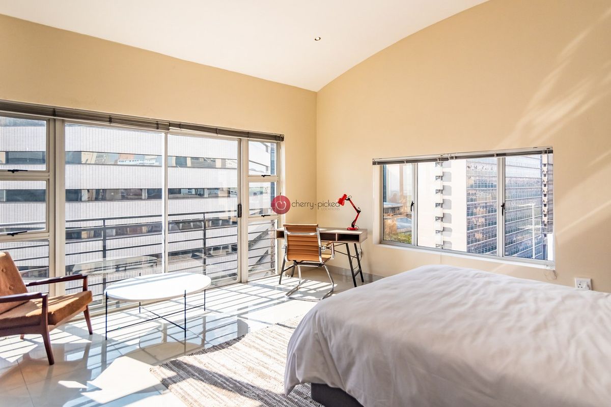 A bright and spacious bedroom featuring high ceilings, large windows, and sliding glass doors that provide ample natural light and urban views.